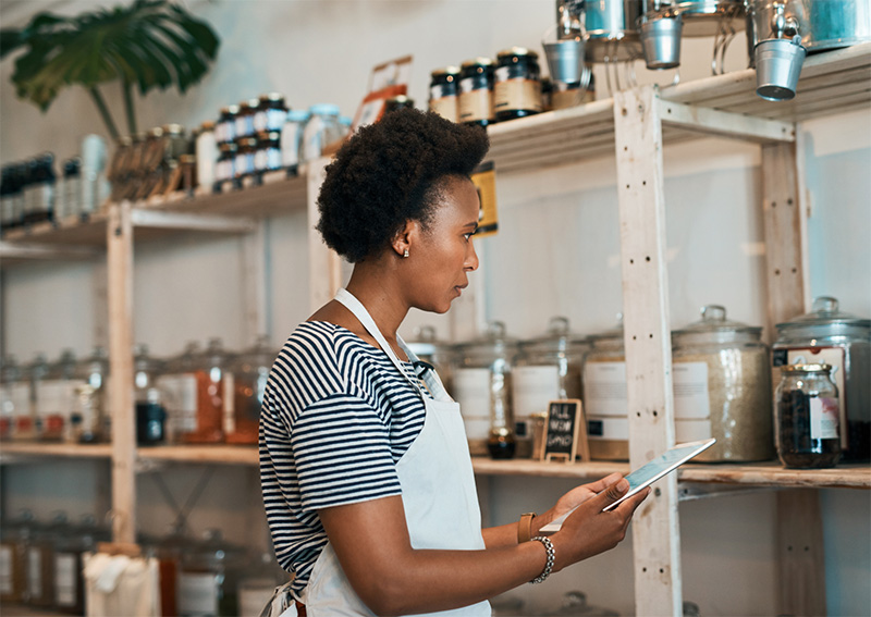 A person wearing an apron stands in a shop with shelves of jars and containers, looking at a digital tablet, possibly checking inventory or managing orders.