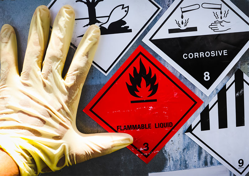 A gloved hand is shown next to warning signs for hazardous materials, including a red flammable liquid sign, a corrosive symbol, and other caution labels.