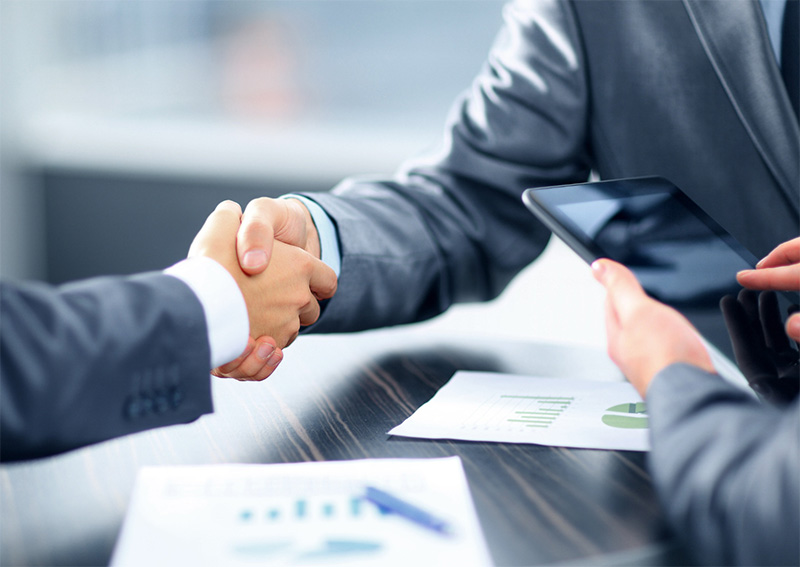 Two people in business suits are shaking hands across a table with documents and a tablet, suggesting a successful business agreement or meeting.