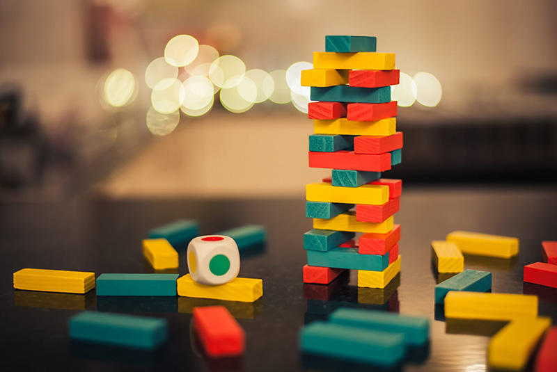A colorful stacking block tower game sits on a table, with scattered blocks and a die nearby. The background is softly blurred with twinkling lights.