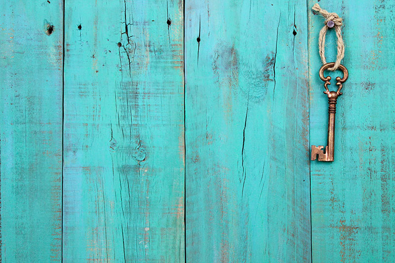 A vintage brass key tied with rope hangs from a nail on a weathered turquoise wooden plank wall.