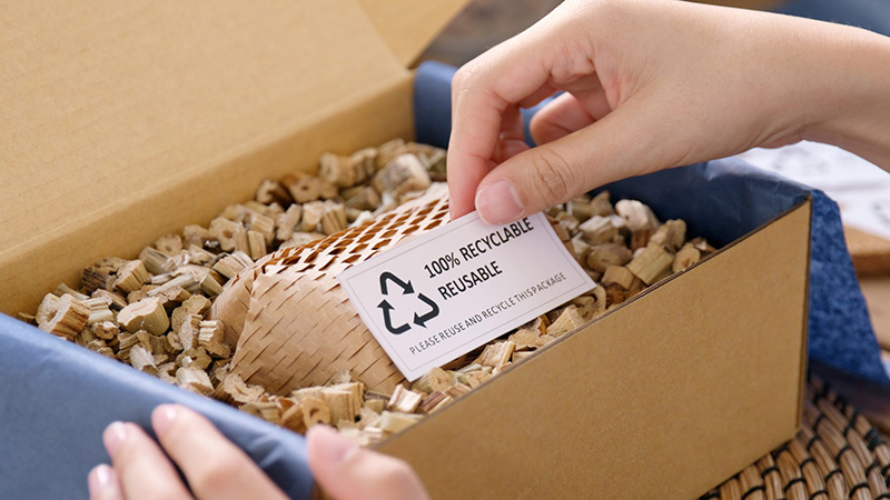 A person opens a cardboard box filled with eco-friendly packing material, revealing a small woven container and a card labeled 100% Recyclable, Reusable with recycling symbols.