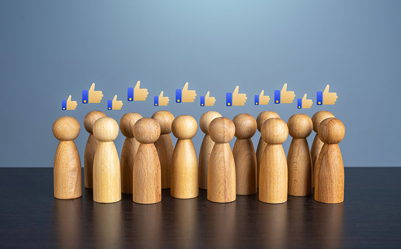 Wooden figurines stand in a group on a dark surface with floating thumbs-up icons above them, symbolizing social approval or popularity. The background is a plain gradient blue.