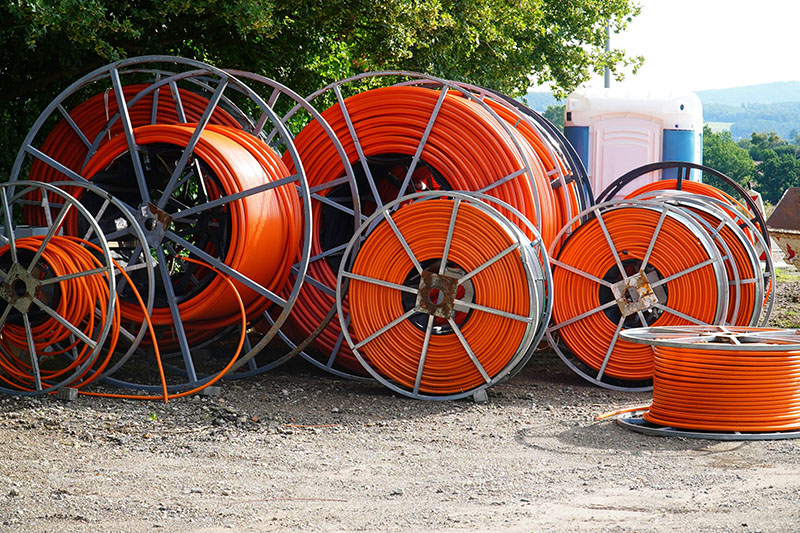 Large metal reels with coiled bright orange cables are arranged outdoors on gravel, with trees, a portable toilet, and hills visible in the background.