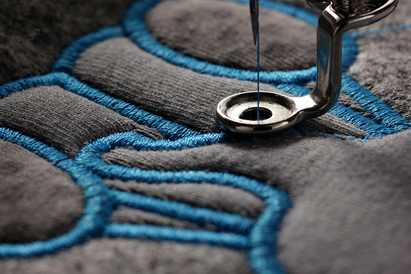 A close-up of a sewing machine needle stitching blue thread into gray fabric, creating an embroidered pattern with smooth curves and bold outlines.