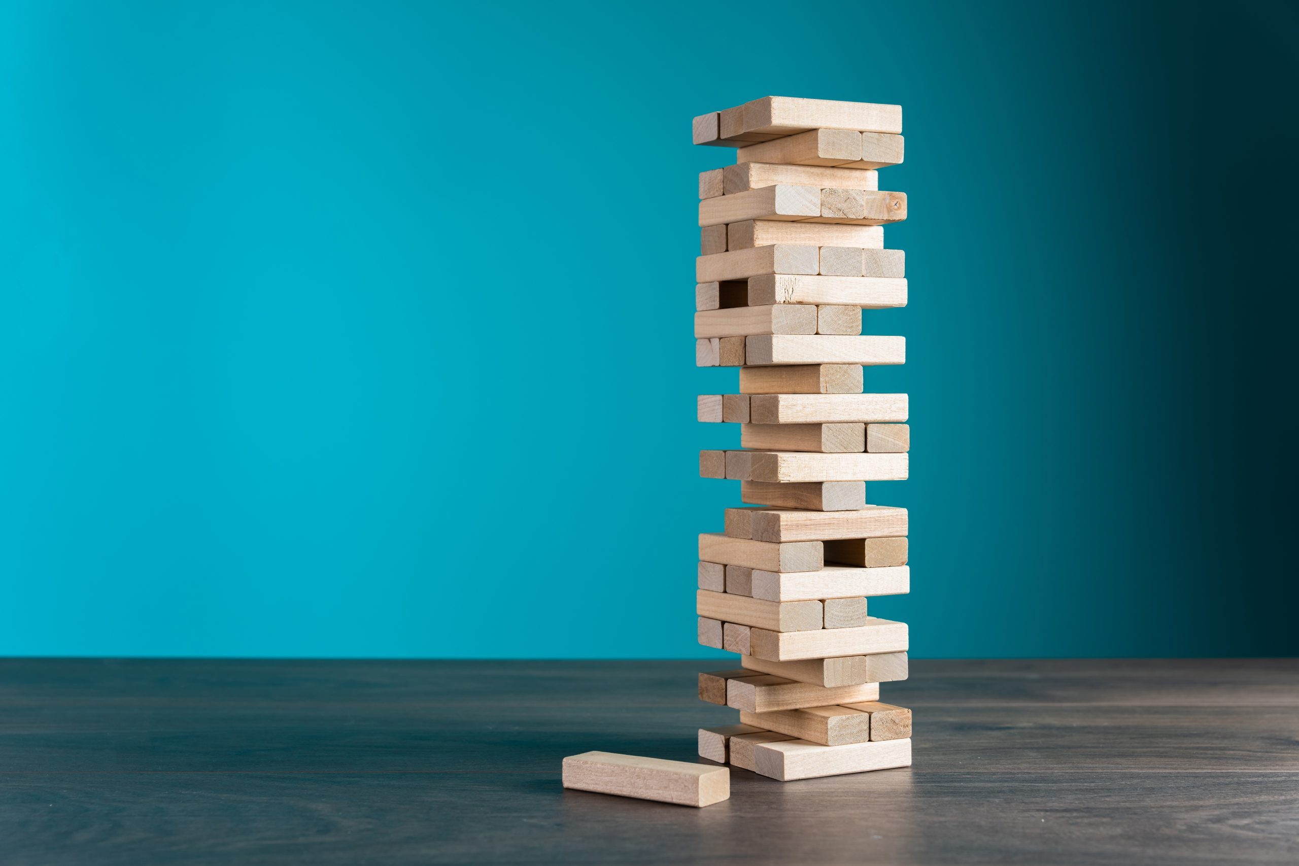 A leaning tower of wooden Jenga blocks on a wooden surface, with one block removed and lying beside the tower, set against a solid teal background.