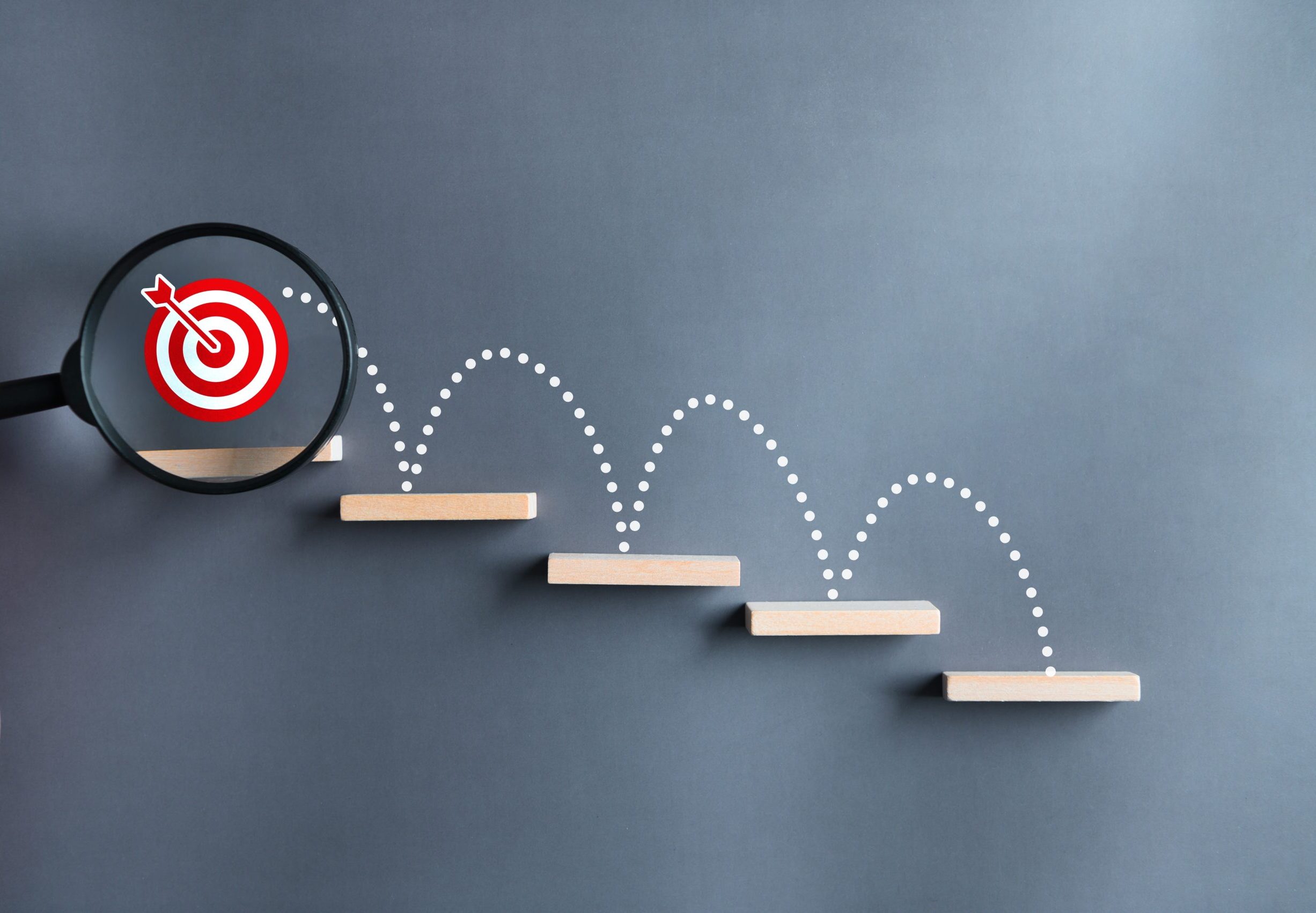 A magnifying glass focuses on a red target symbol at the top of a staircase made of wooden steps, with a dotted line illustrating a path upward over each step.