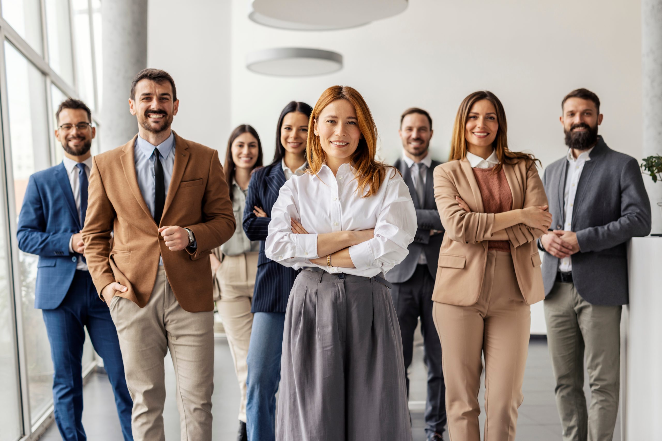 A group of eight professionally dressed people stand together in a bright office, smiling confidently at the camera. The woman in front has her arms crossed, with colleagues standing behind her in business attire.