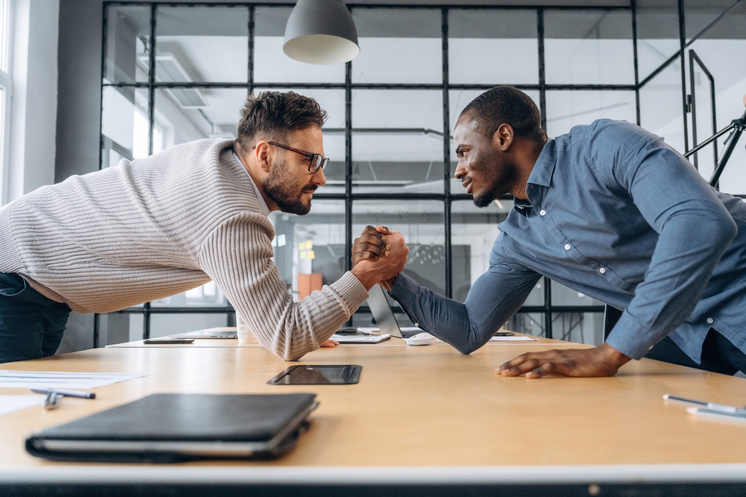 Two men arm wrestling across a table in a modern office setting, both smiling and appearing competitive. Laptops and office supplies are on the table, and glass partitions are in the background.